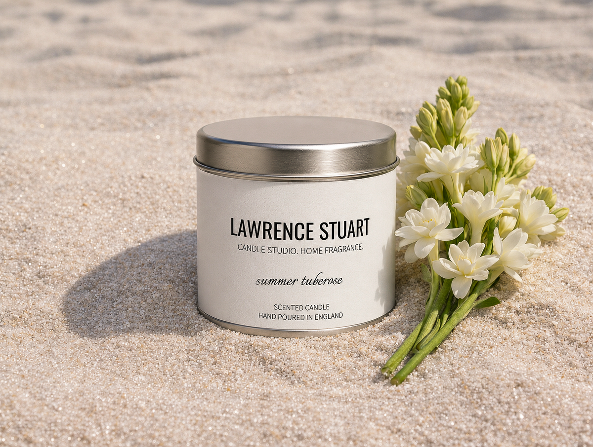 A Summer Tuberose candle tin displayed in beach sand beside a bouquet of tuberose flowers.